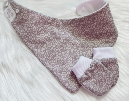A baby bib and matching booties set in a dusty lavender and white floral pattern, laid flat on a white faux fur background.