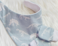 A close-up of a baby bandana bib in light blue with a white skeleton dinosaur pattern, laid flat on a white faux fur surface.