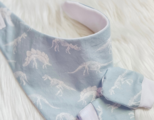 A close-up of a baby bandana bib in light blue with a white skeleton dinosaur pattern, laid flat on a white faux fur surface.
