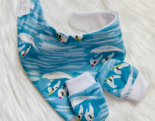 A baby clothing set including a bib and matching booties, featuring a light blue and white watercolor wave pattern with penguin and surfers printed across the fabric, displayed on a white faux fur background.