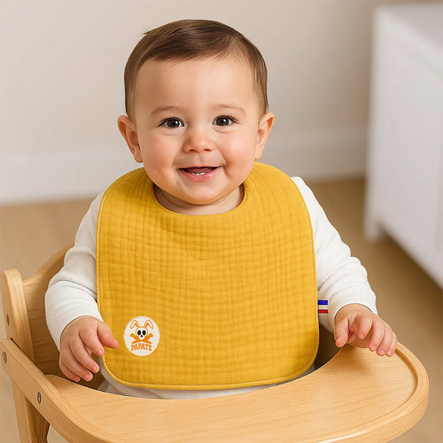 Baby wearing a yellow bib with a logo, sitting in a wooden high chair.