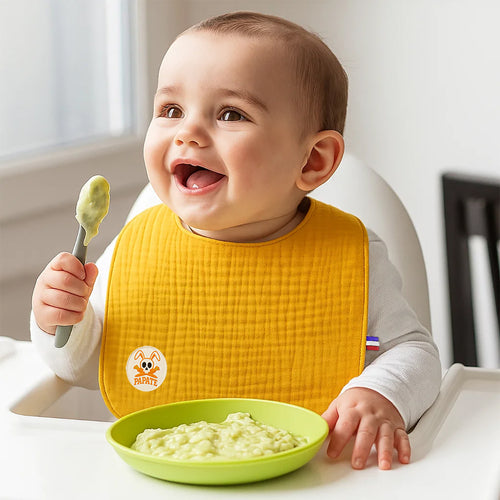 Baby in a high chair with a yellow bib, holding a spoon and a green bowl with food.